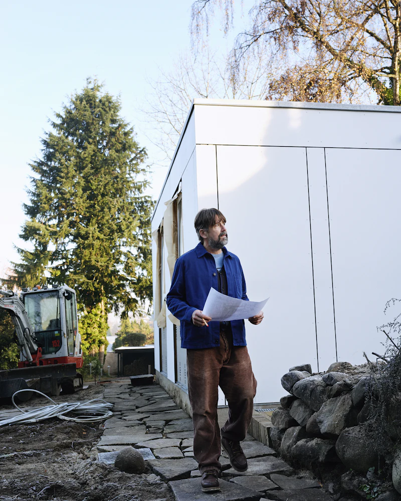 Ein Bauleiter in blauer Arbeitsjacke liest vor einem Neubau einen Plan, hinter ihm steht ein Minibagger.