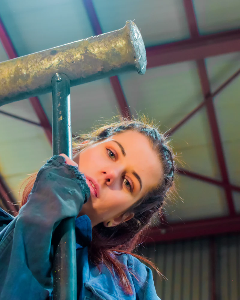 Eine Handwerkerin im blauen Arbeitsanzug stützt sich in einer Halle auf einen schweren Hammer.