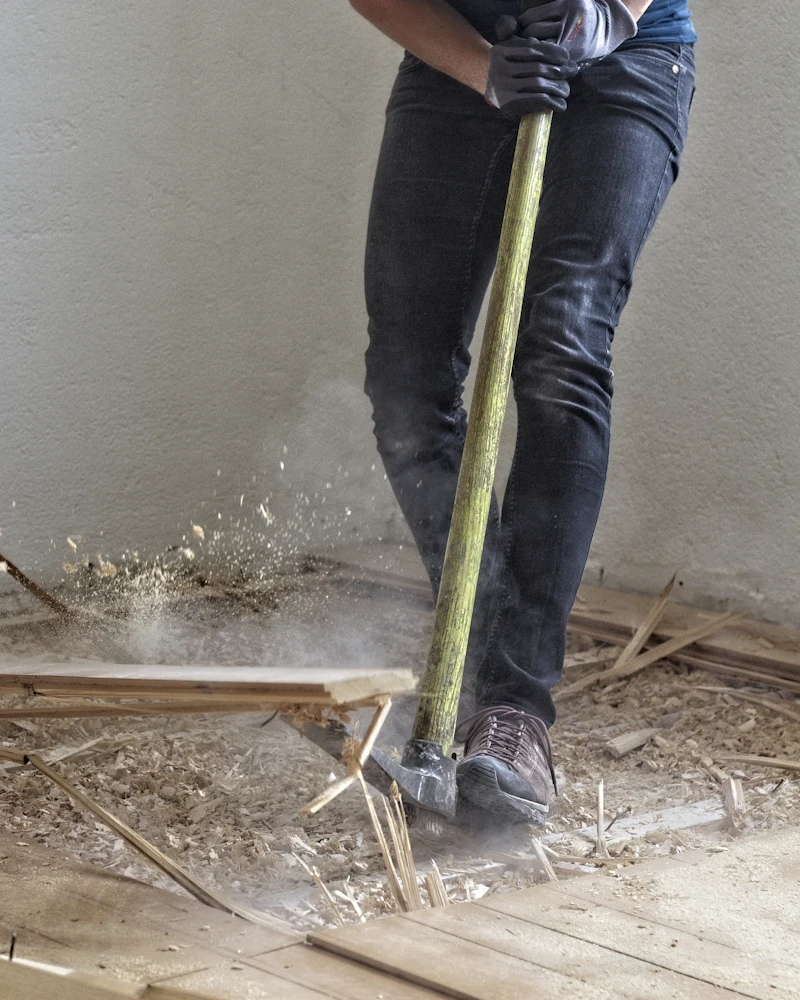 Ein Heimwerker in Jeans und Arbeitshandschuhen reißt mit dem Vorschlaghammer einen alten Holzboden heraus.